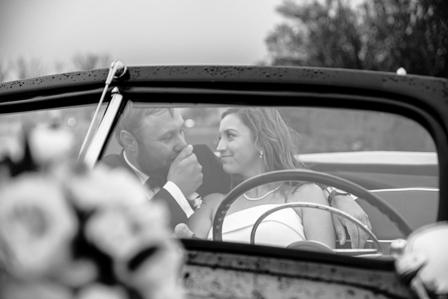 Black and white photo of a bride and groom sharing a tender moment inside a vintage classic car on their wedding day.