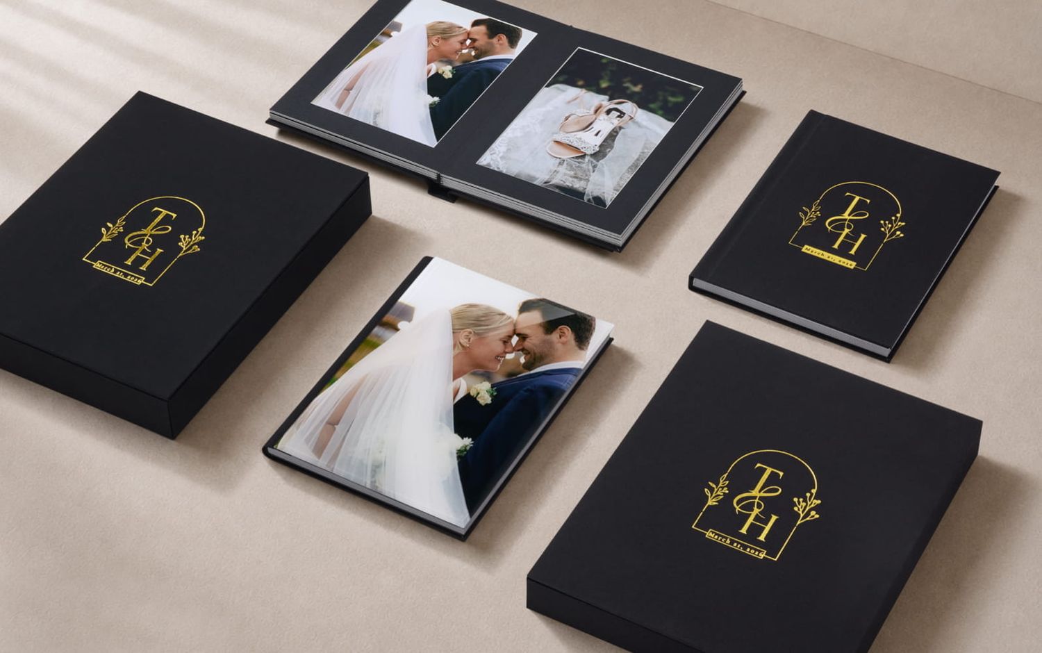 Black gold-foil monogram wedding album boxes with open photo albums showing bridal couple portraits on a neutral tabletop.