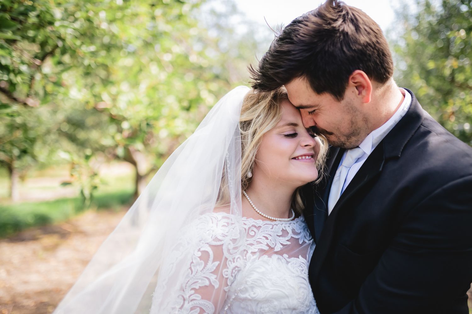 A tender moment shared between newlyweds in lace and formal attire outdoors with greenery in background.