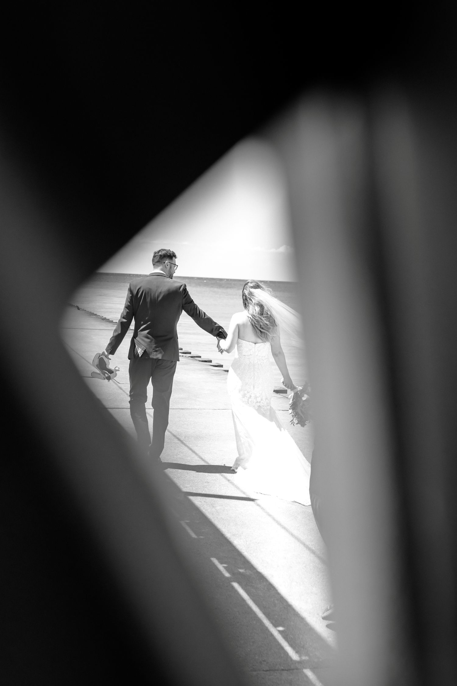 Black and white photo of bride and groom holding hands, viewed through a gap, walking in sunlight.