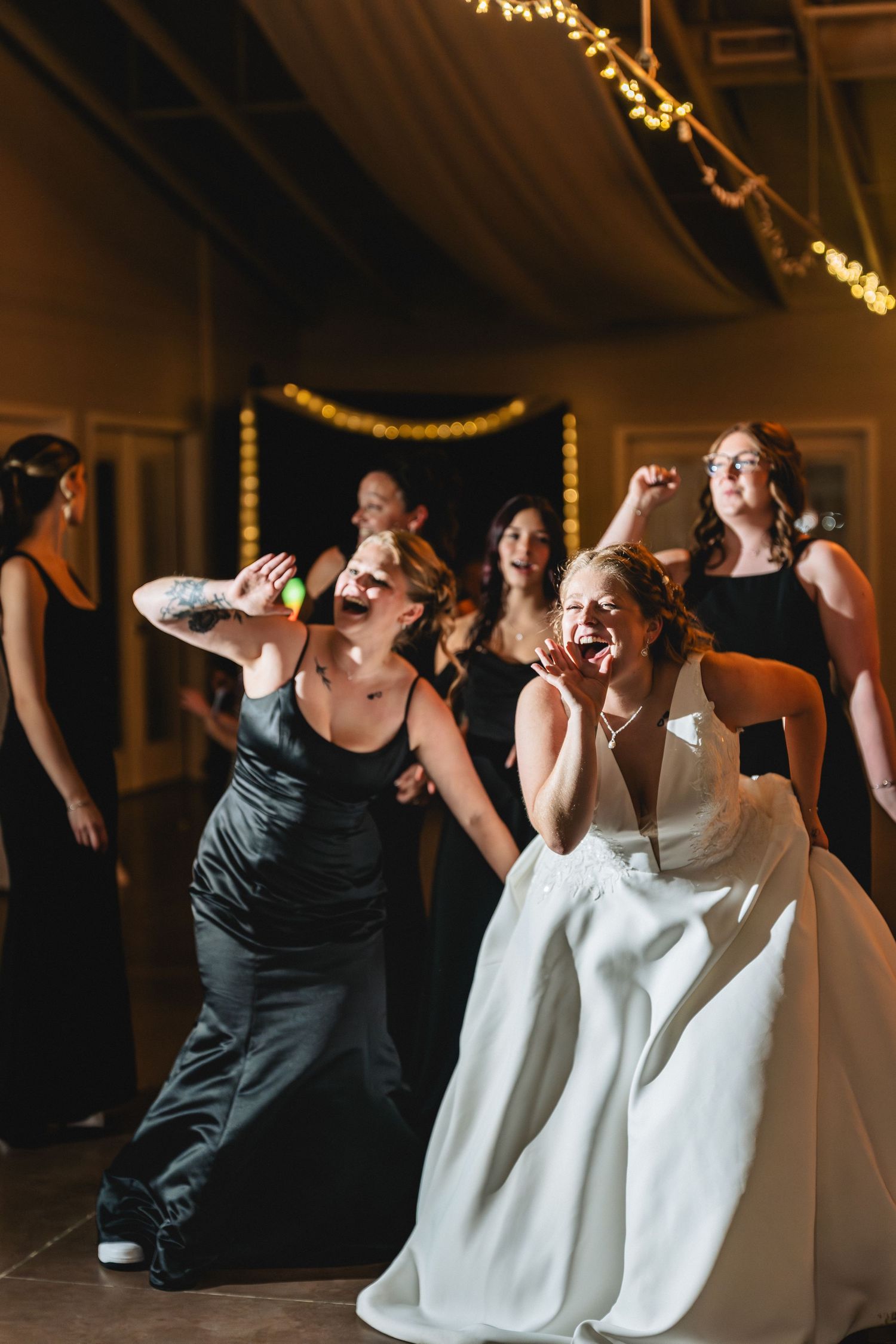 Bride in white gown laughs joyfully on dance floor with bridesmaids in black dresses at wedding reception.