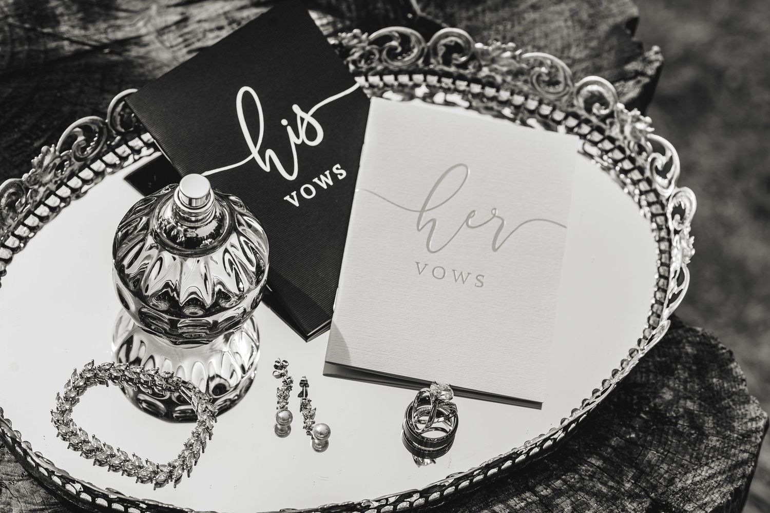 Black and white photo of his and her wedding vow books on a mirrored tray with jewelry and a decorative jar captured in Traverse City