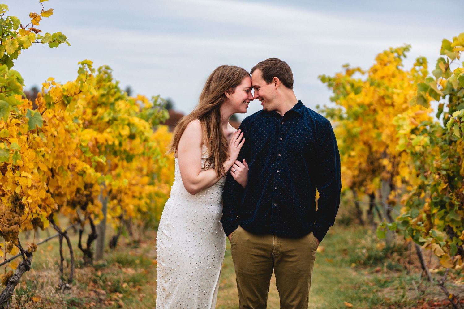 Couple sharing a romantic moment in a golden autumn vineyard, wearing white dress and navy shirt.