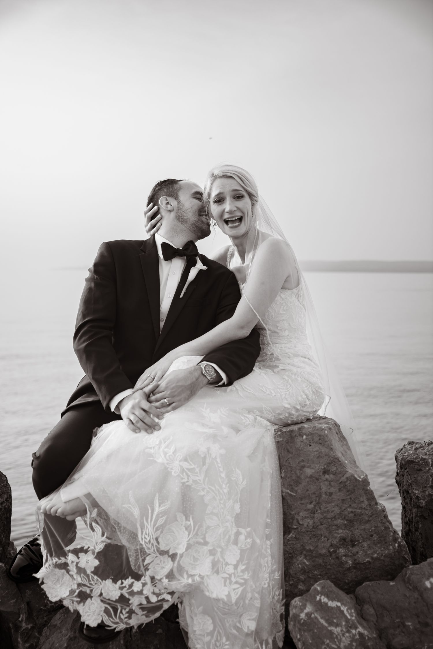 Black and white photo of a groom kissing bride on cheek as she laughs joyfully, sitting on rocks by the water.