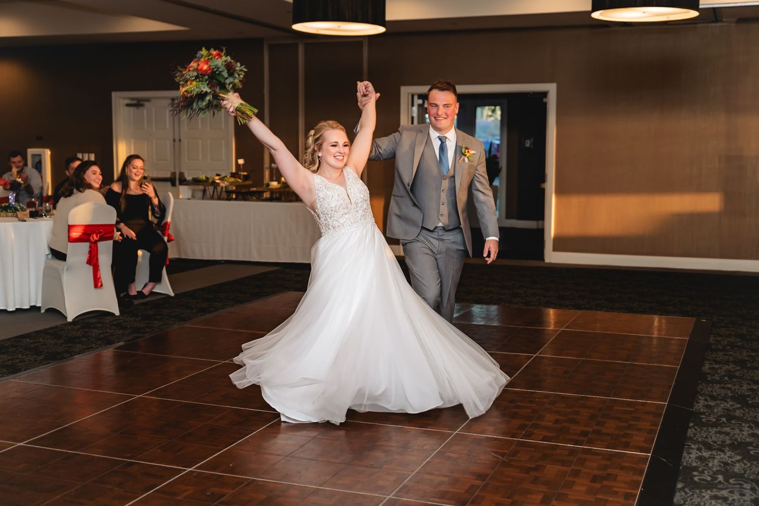 A couple performs their first dance on a dark wooden floor at a wedding reception venue with guests watching.