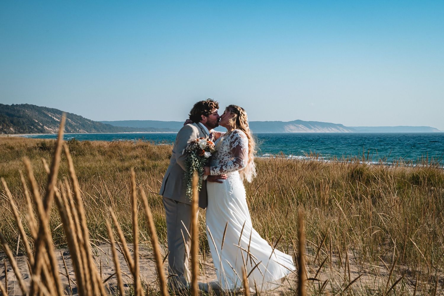A couple shares a romantic moment on a coastal path with tall grass and ocean views in the background.