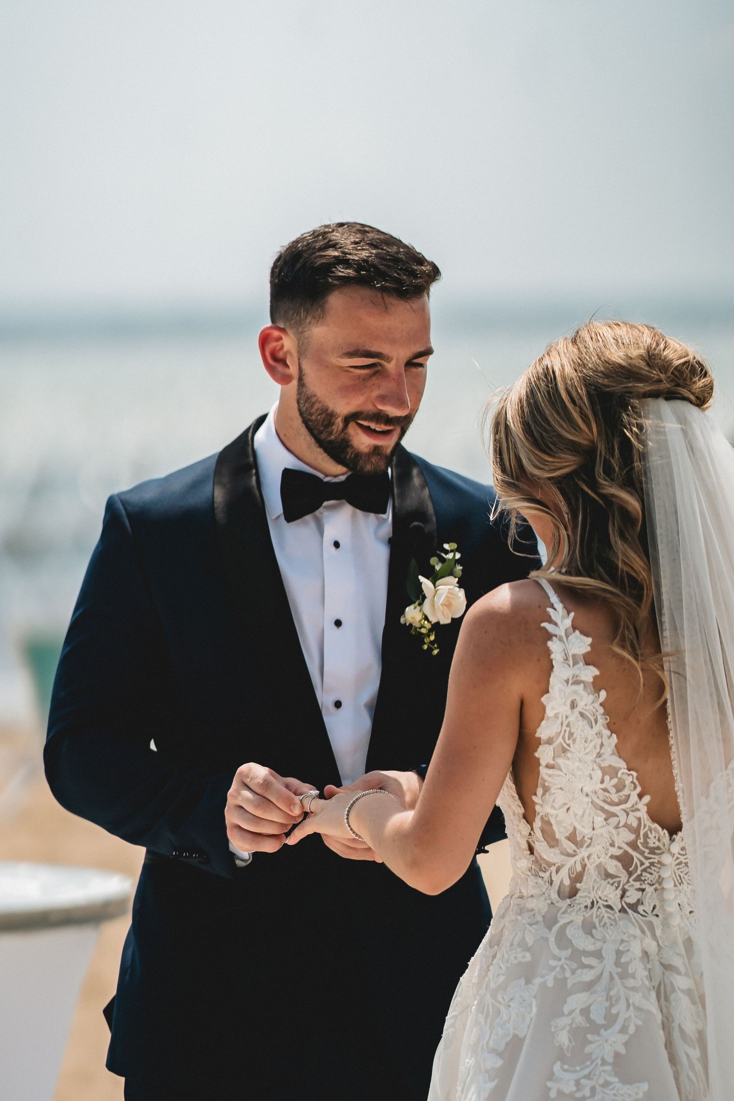 Groom in black tuxedo placing wedding ring on bride in lace dress outdoors near water.