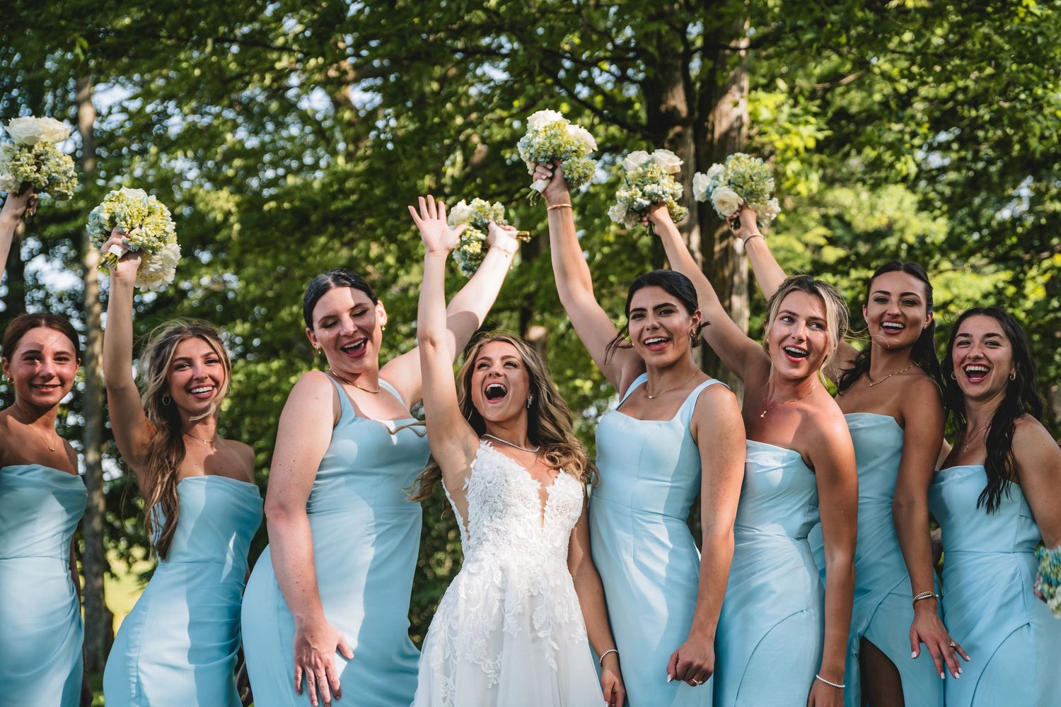 Bride in white gown celebrates with bridesmaids in light blue dresses, holding bouquets outdoors among lush green trees.