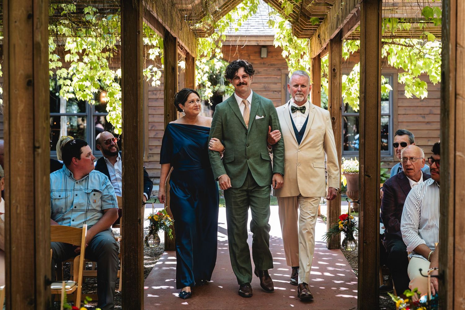 Groom walks down the aisle with two family members at an outdoor wedding ceremony under a vine-covered pergola.