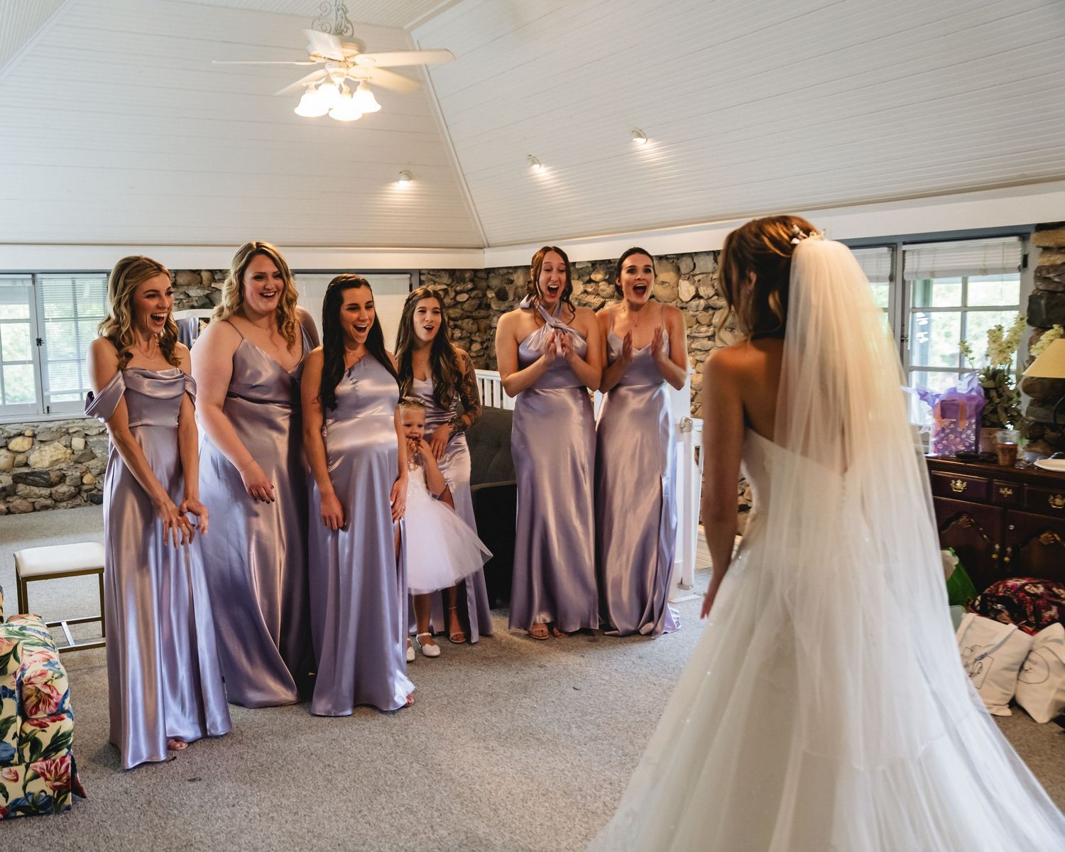 Bride reveals wedding dress to bridesmaids in lavender gowns inside a rustic stone chapel venue.