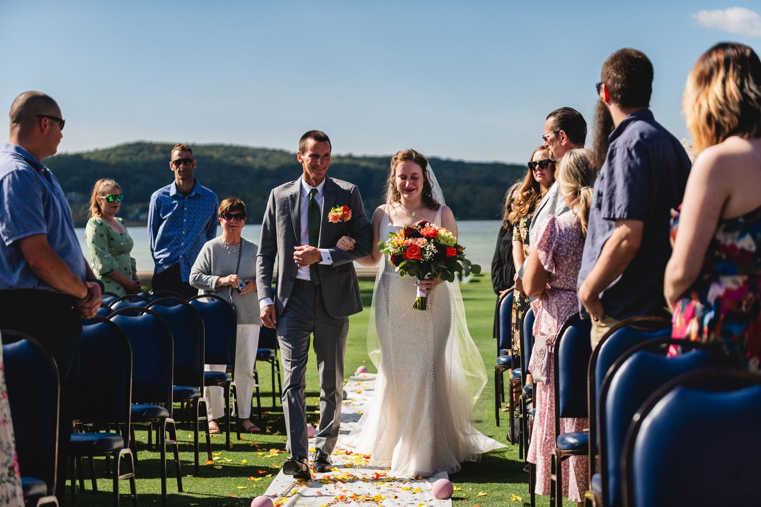 Wedding guests gather outdoors for a scenic ceremony with mountains and water views in the background.
