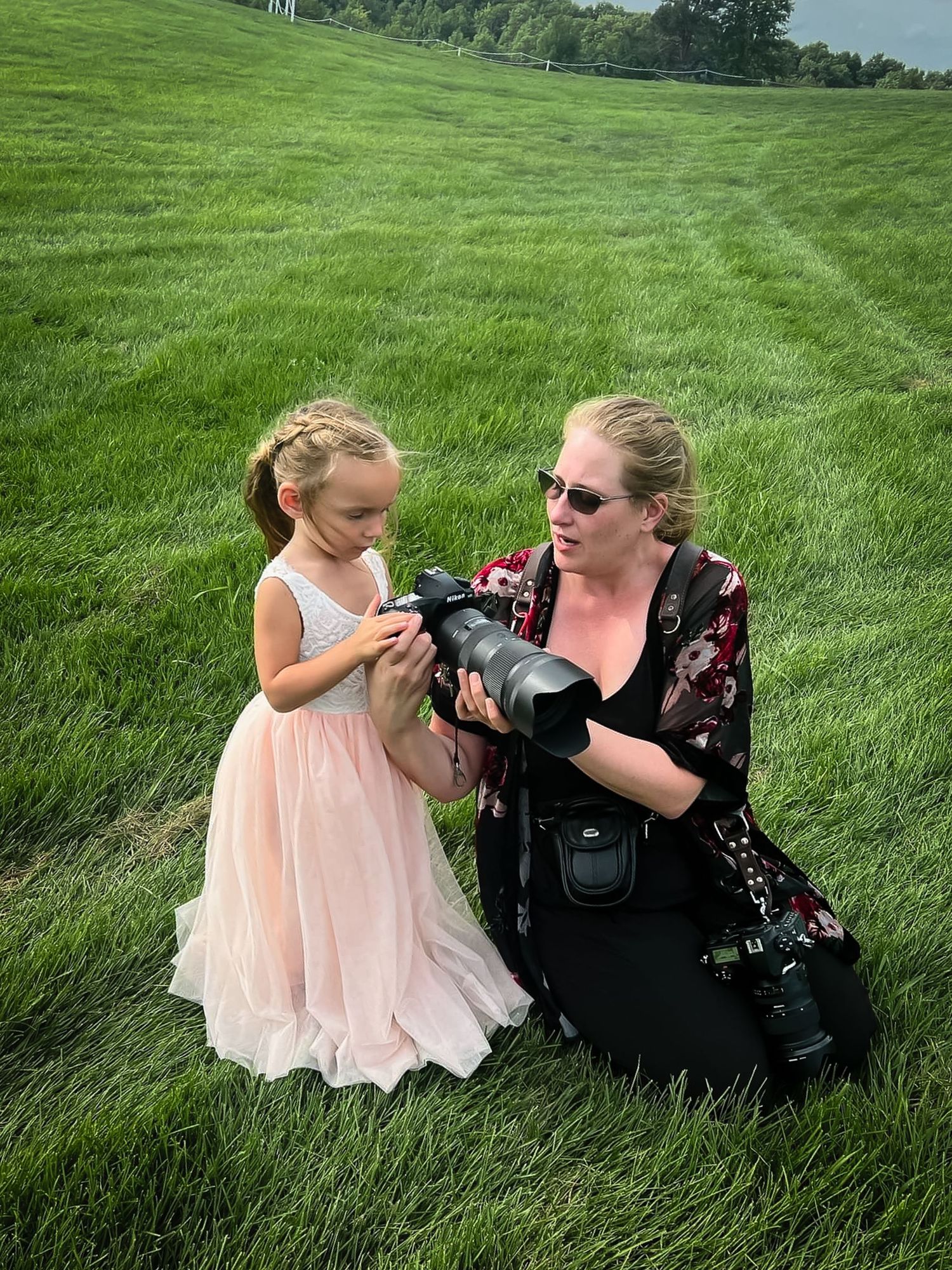 A woman photographer shows a young girl in a pink dress her camera on a green grass field outdoors.
