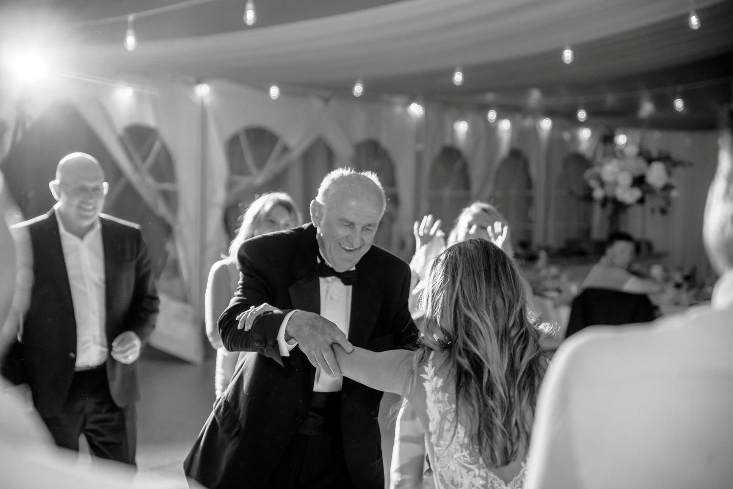 Black and white photo of elderly man dancing with bride at elegant wedding reception under string lights.