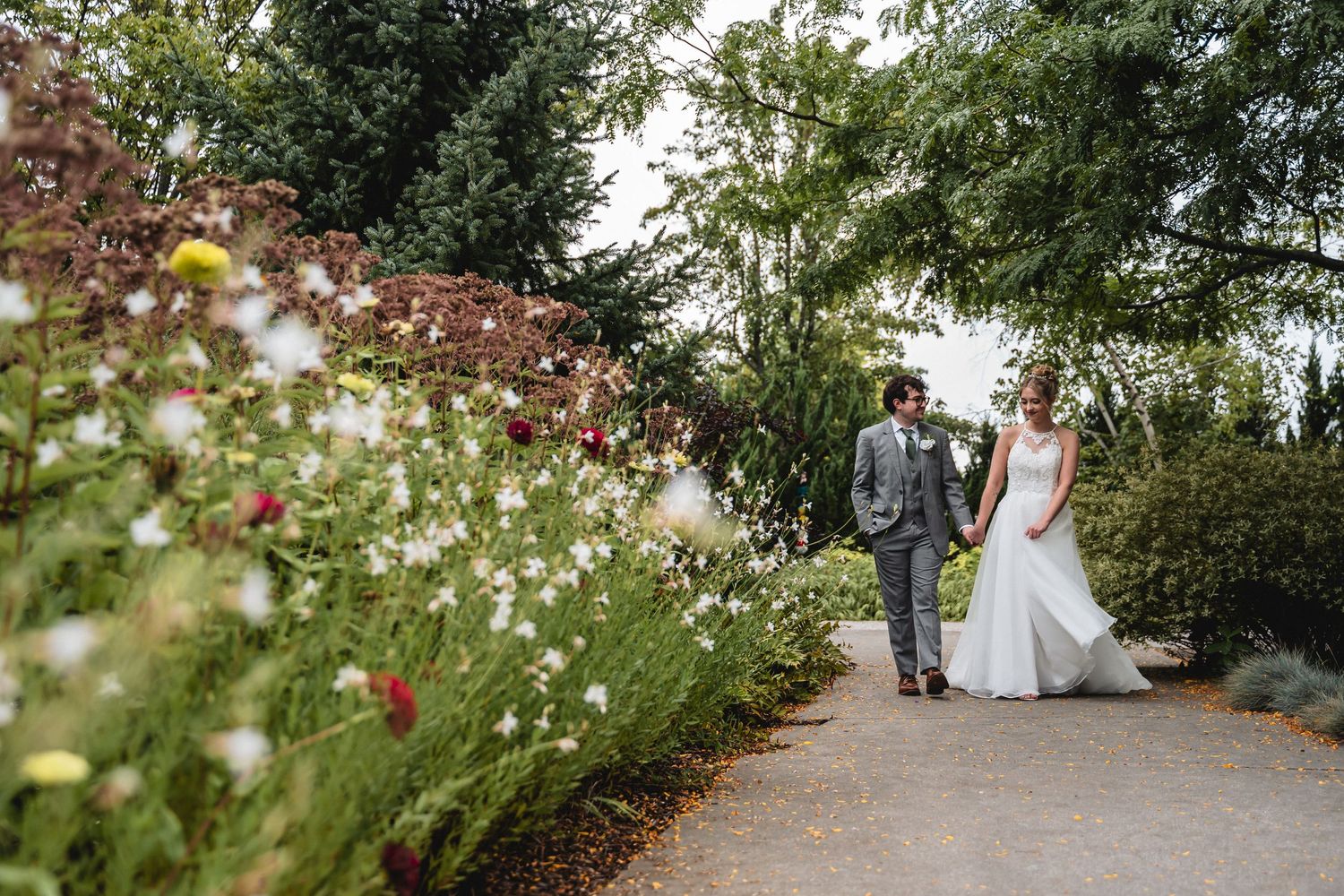 Bride and groom walk hand in hand along a garden path surrounded by colorful wildflowers on their wedding day.