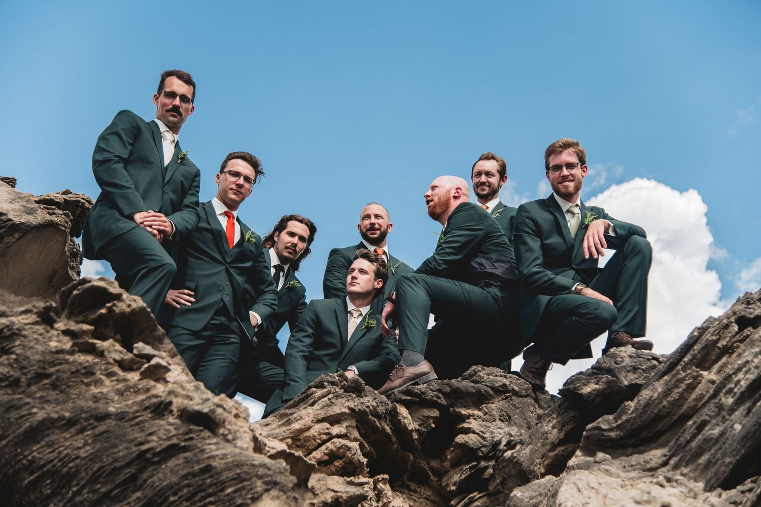 Eight men in matching dark green suits pose on rocky terrain under a blue sky, shot from a low angle.