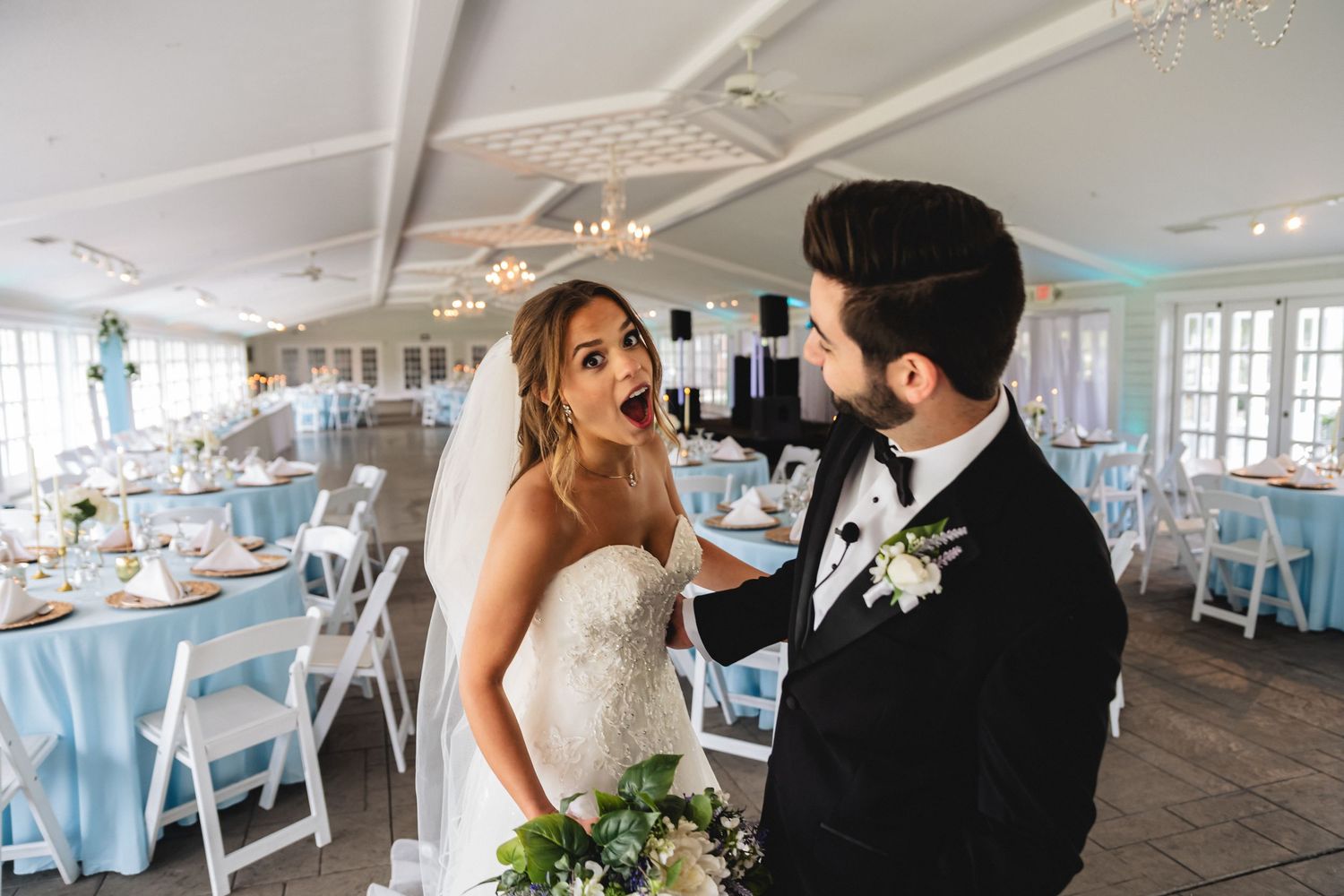Wedding reception tables decorated in elegant blue and white with floral centerpieces in a bright sunlit venue.