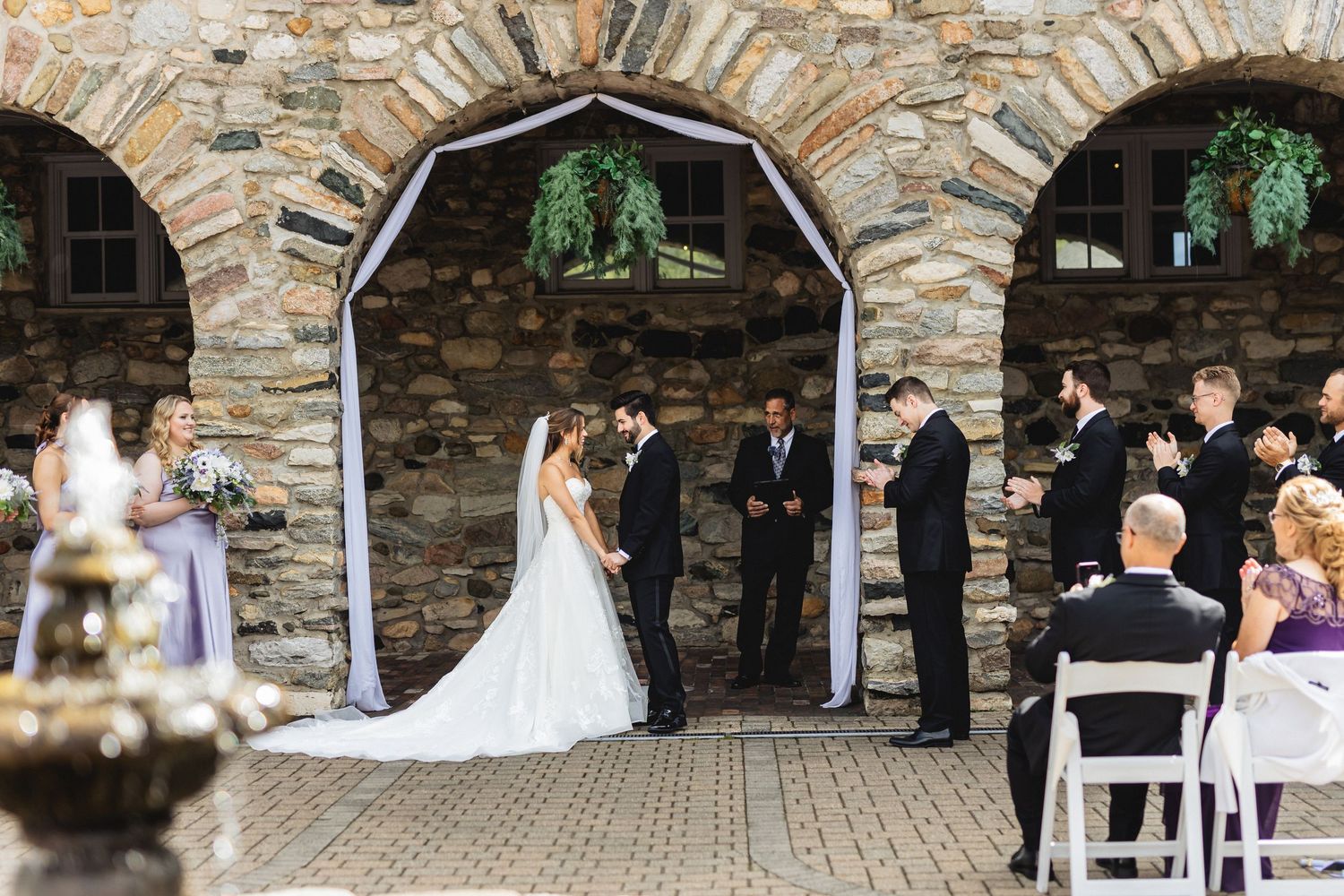 Wedding ceremony under stone archway with guests seated in white chairs during outdoor celebration.