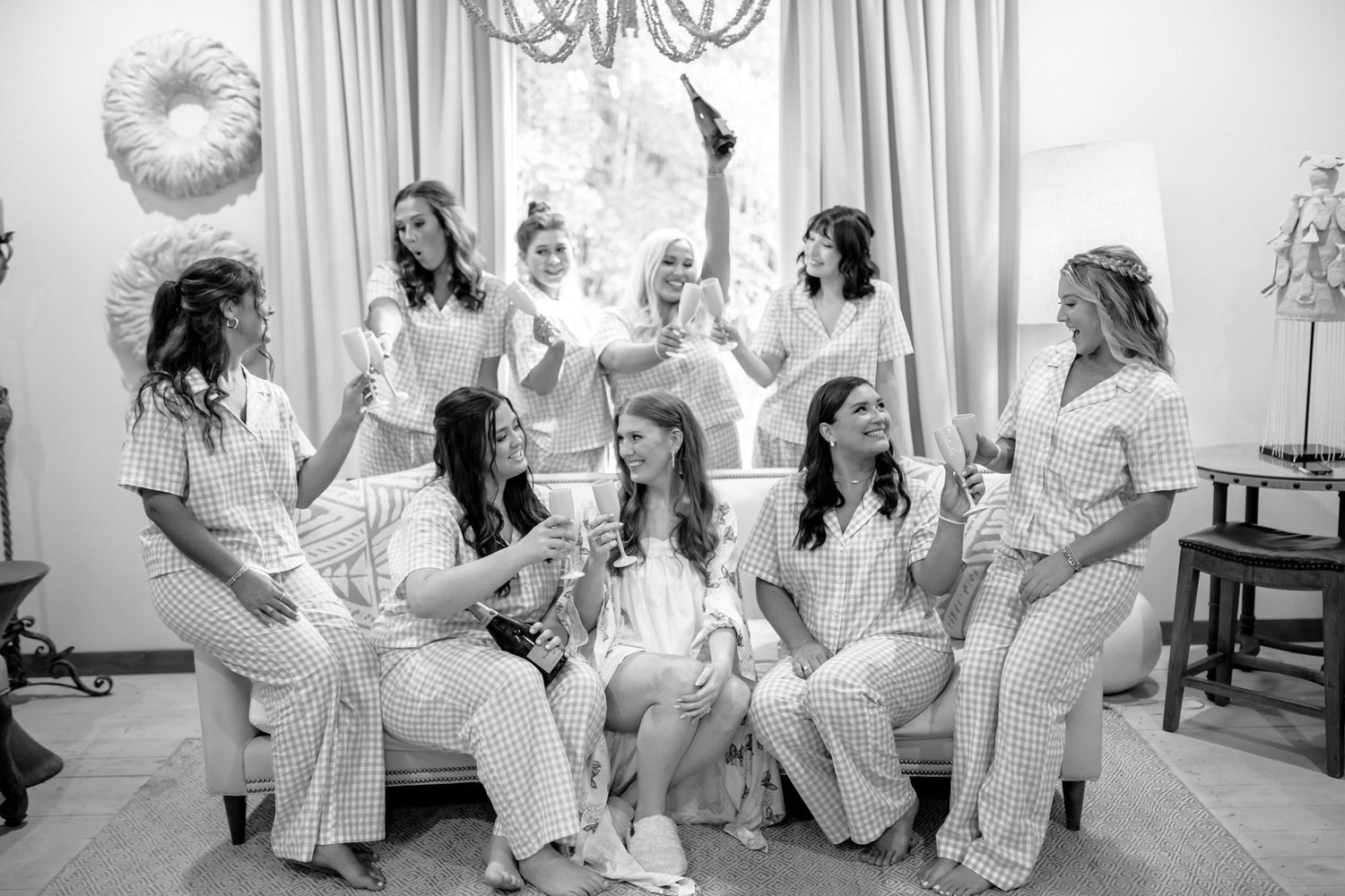Bridesmaids in matching striped pajamas laugh and toast with champagne while gathered on a sofa in a bright room before the wedding.