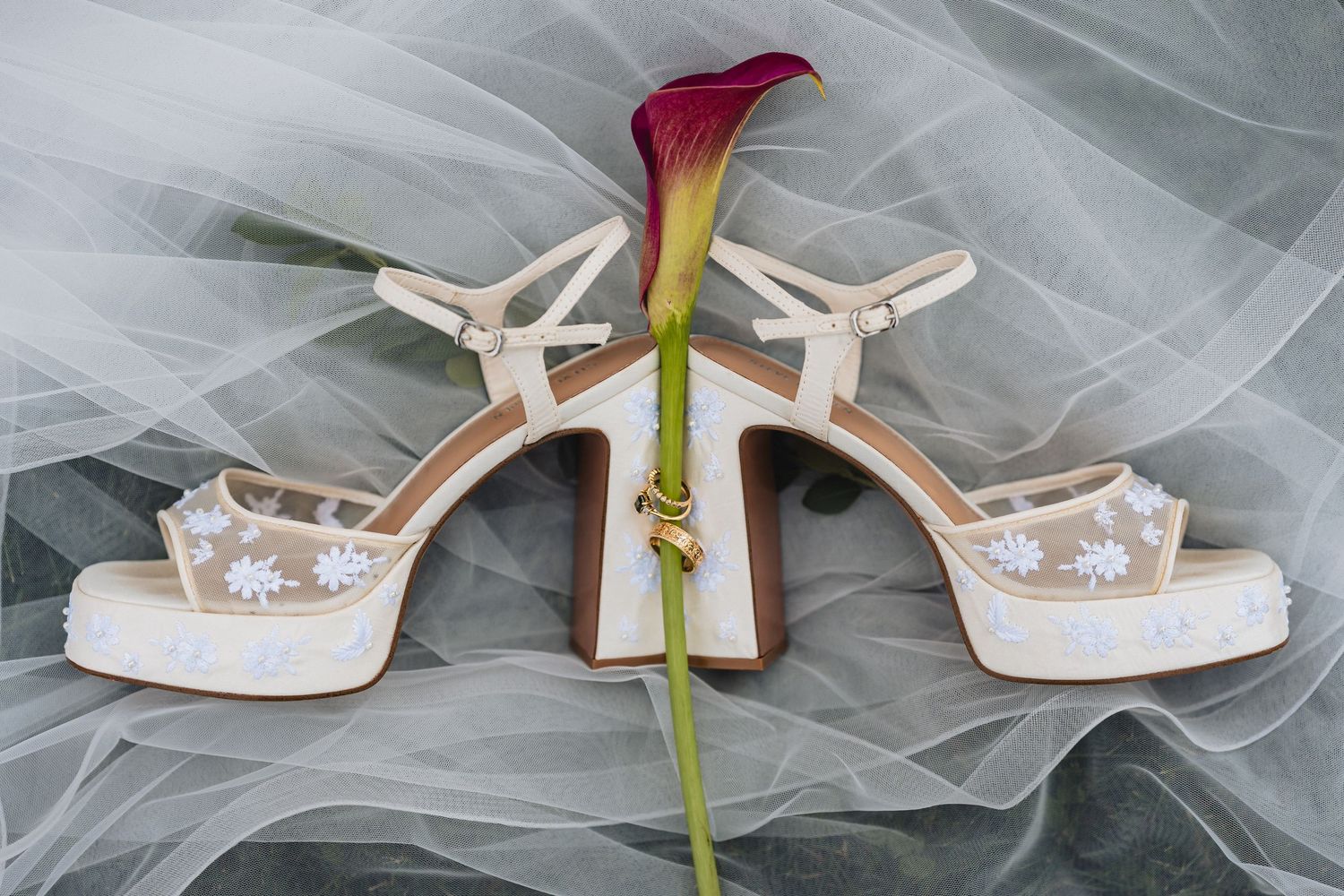 Elegant bridal platform sandals with floral details rest on sheer veil beside a burgundy calla lily.