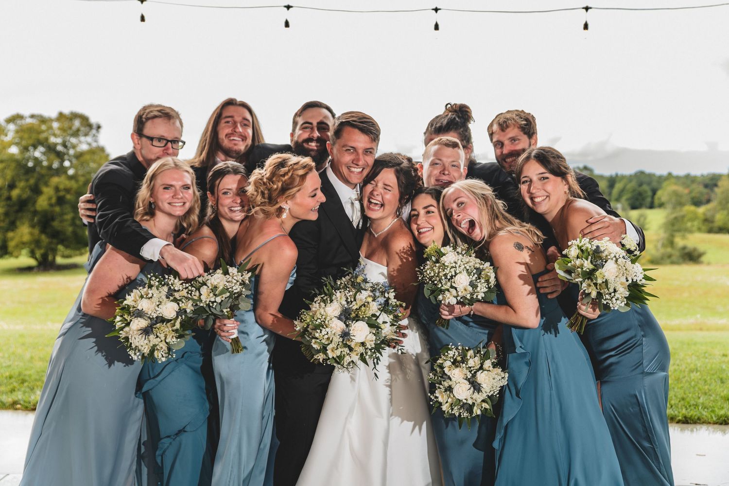 Wedding party in blue dresses and black suits surrounds smiling bride and groom outdoors.