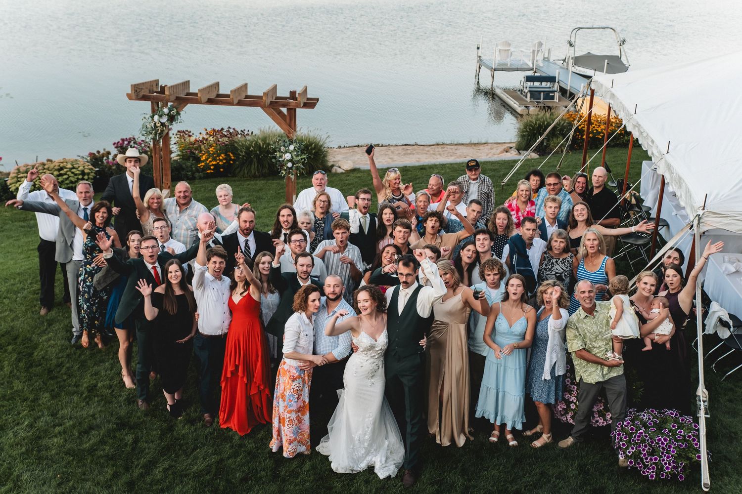 Large wedding group photo outdoors by waterfront with bride and groom centered among smiling guests near a wooden arch.