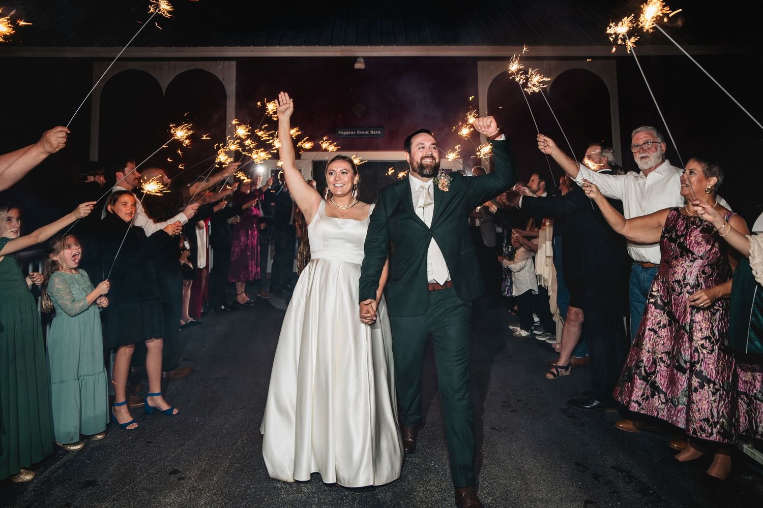 Bride and groom walk through sparkler exit surrounded by smiling wedding guests at night.