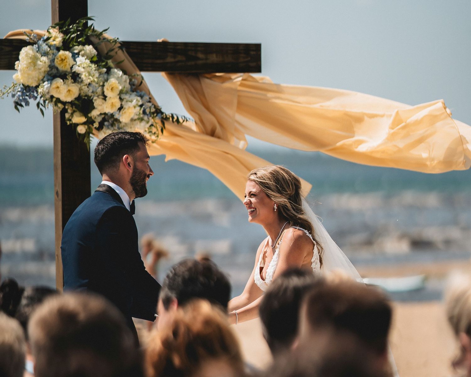 Couple exchanging vows at outdoor beach wedding ceremony under wooden cross arch with yellow fabric and floral decor.