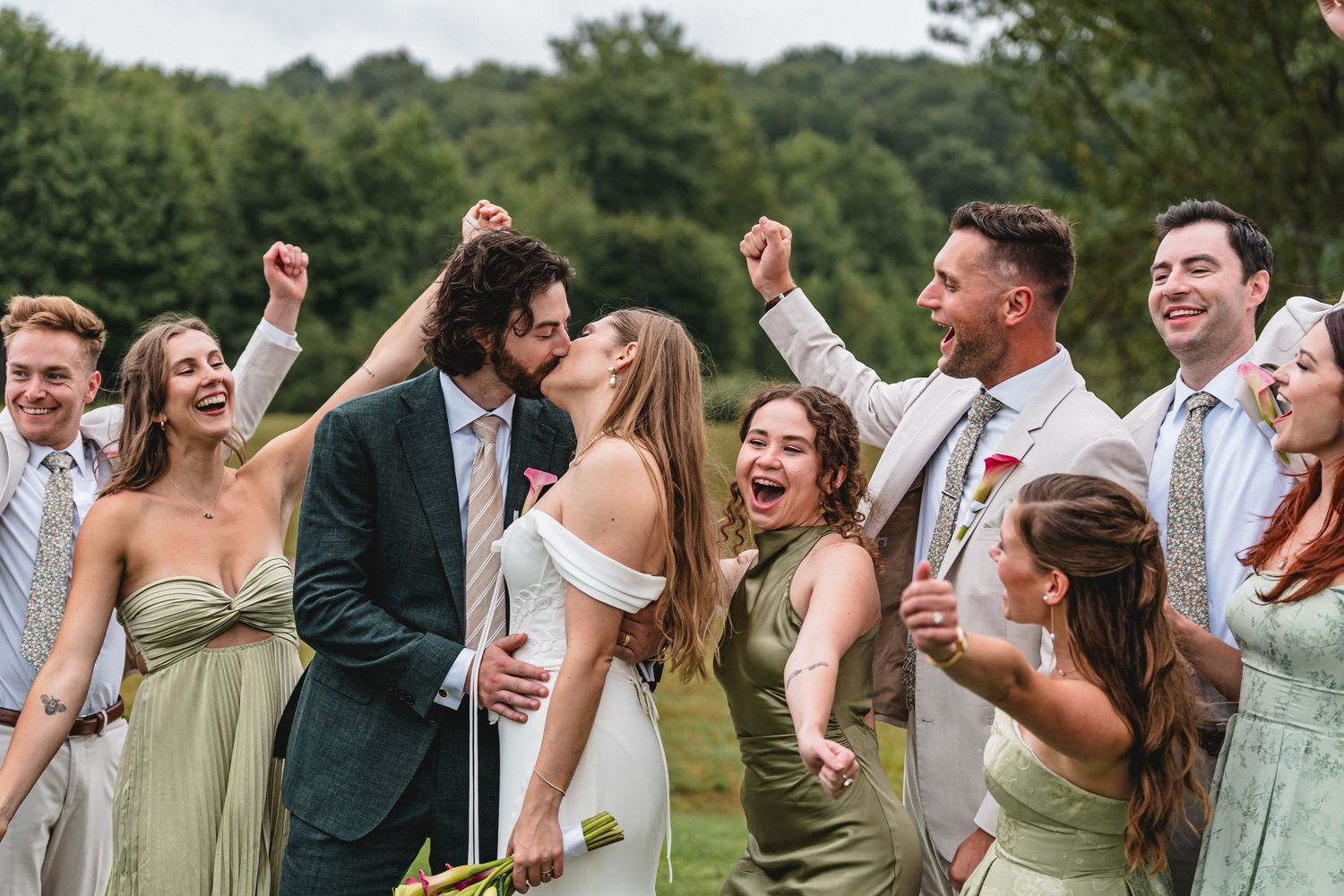 Bride and groom share a kiss outdoors while cheering bridesmaids and groomsmen celebrate around them at The Cherry Barn at Nugent Orchards