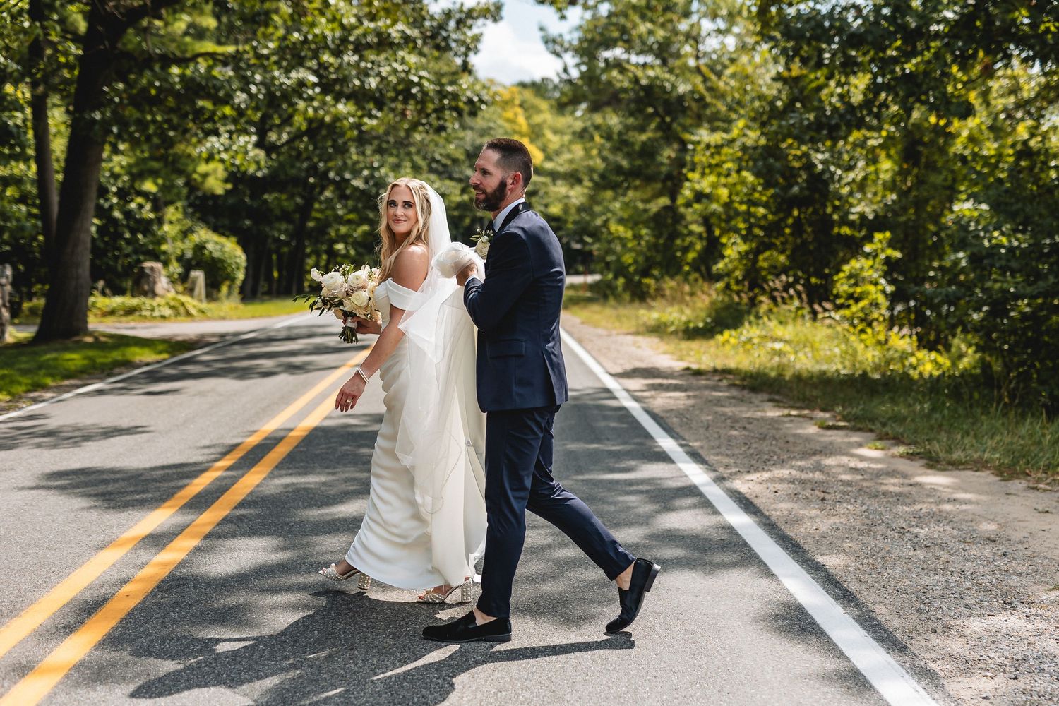 Bride holding a bouquet walks hand in hand with a groom in a navy suit across a tree-lined road on their wedding day at The Peninsula Room