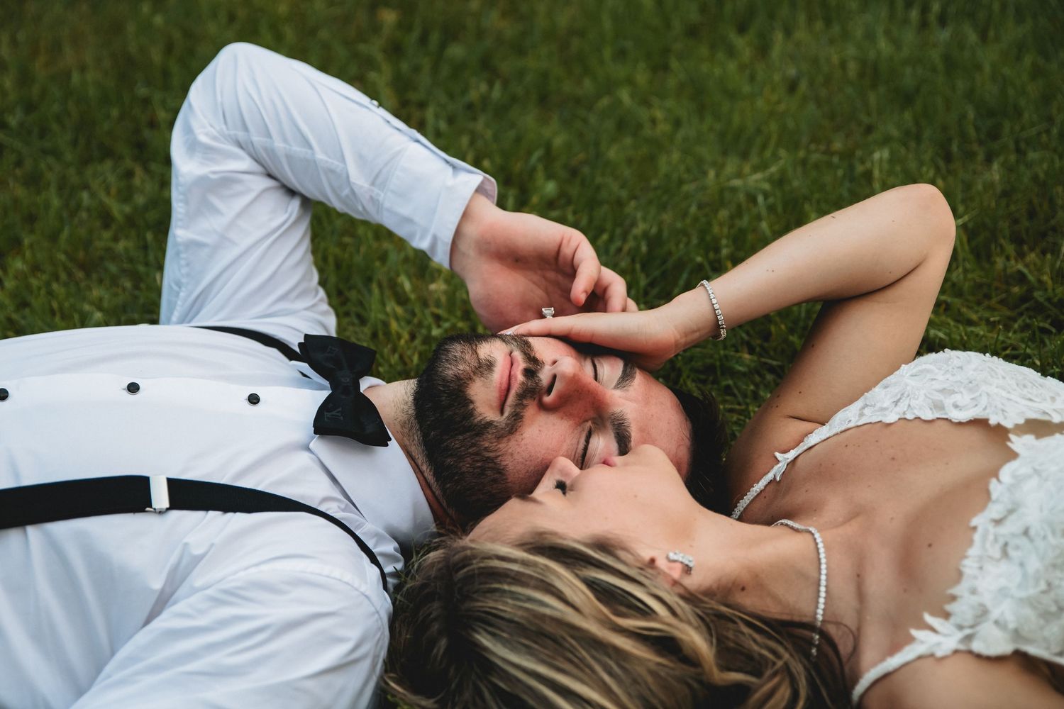 A bride and groom lying on green grass, smiling and gazing at each other on their wedding day.