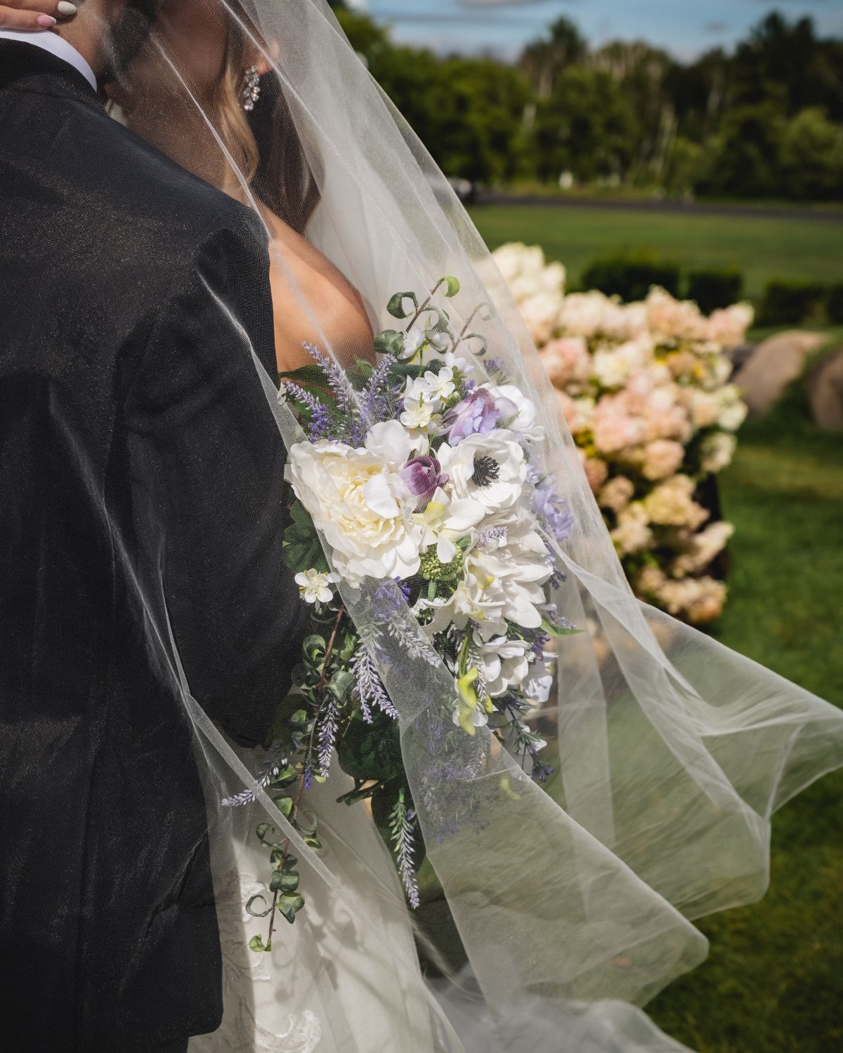 Delicate white and purple floral wedding bouquet with flowing veil detail against outdoor garden backdrop.