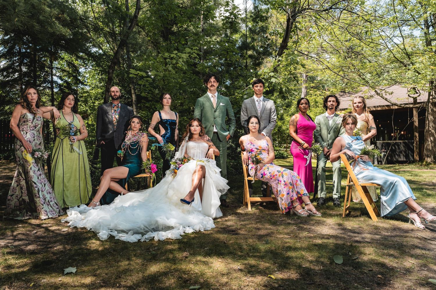 Wedding party poses outdoors in a lush garden setting, featuring bride in white gown surrounded by elegantly dressed guests.
