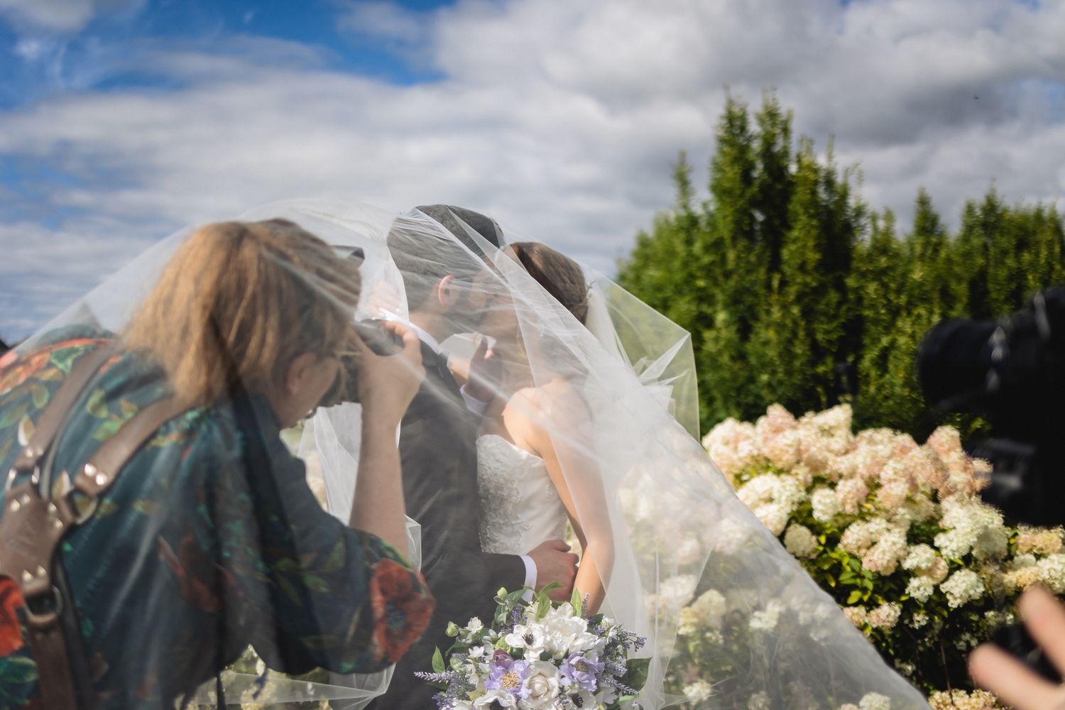 Bride in white veil with wedding guests outdoors, surrounded by white hydrangeas under a blue cloudy sky.