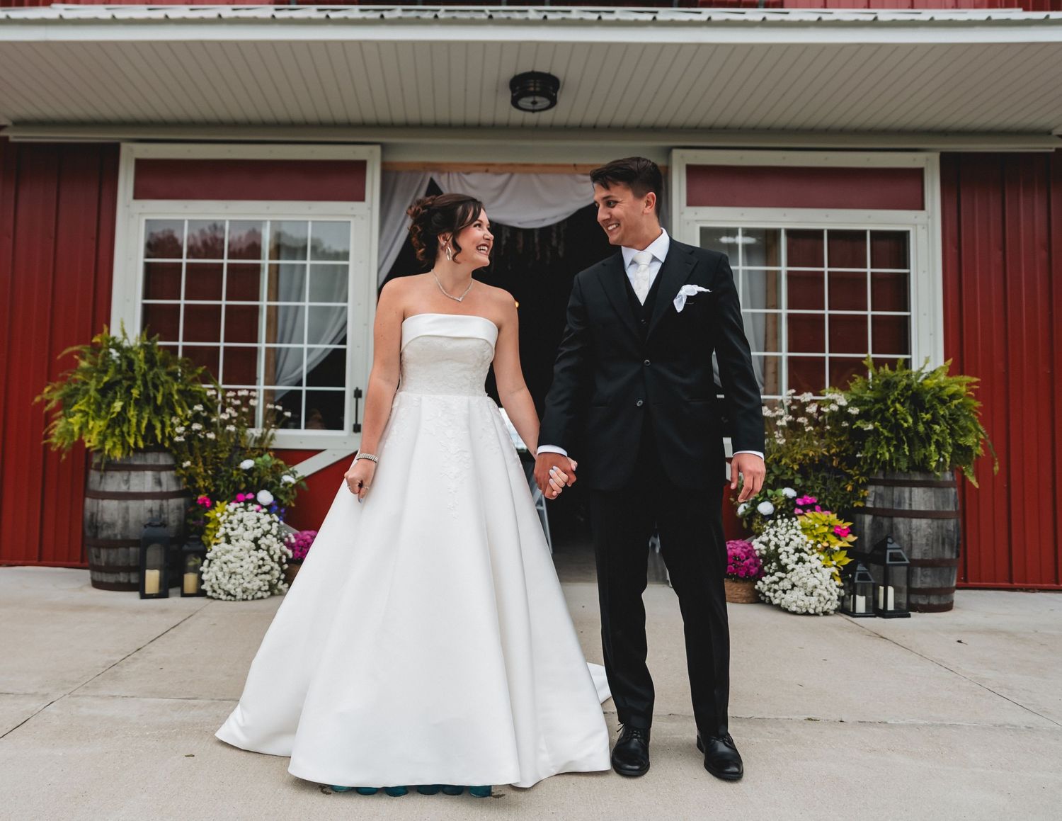 Elegant wedding couple in formal attire stands outside a red barn venue with colorful floral arrangements.