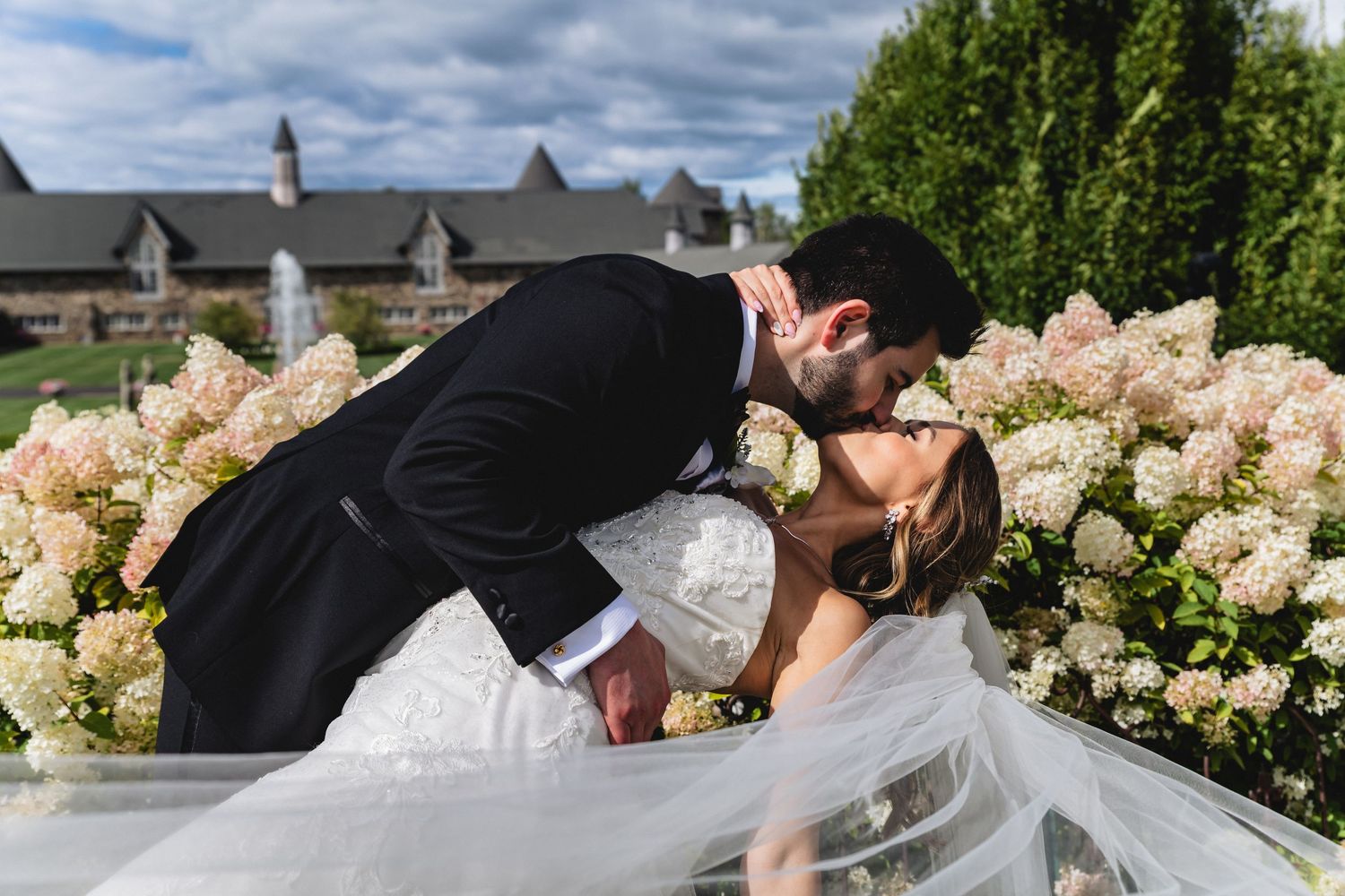 Romantic wedding couple sharing a tender kiss outdoors surrounded by blooming white hydrangea flowers.