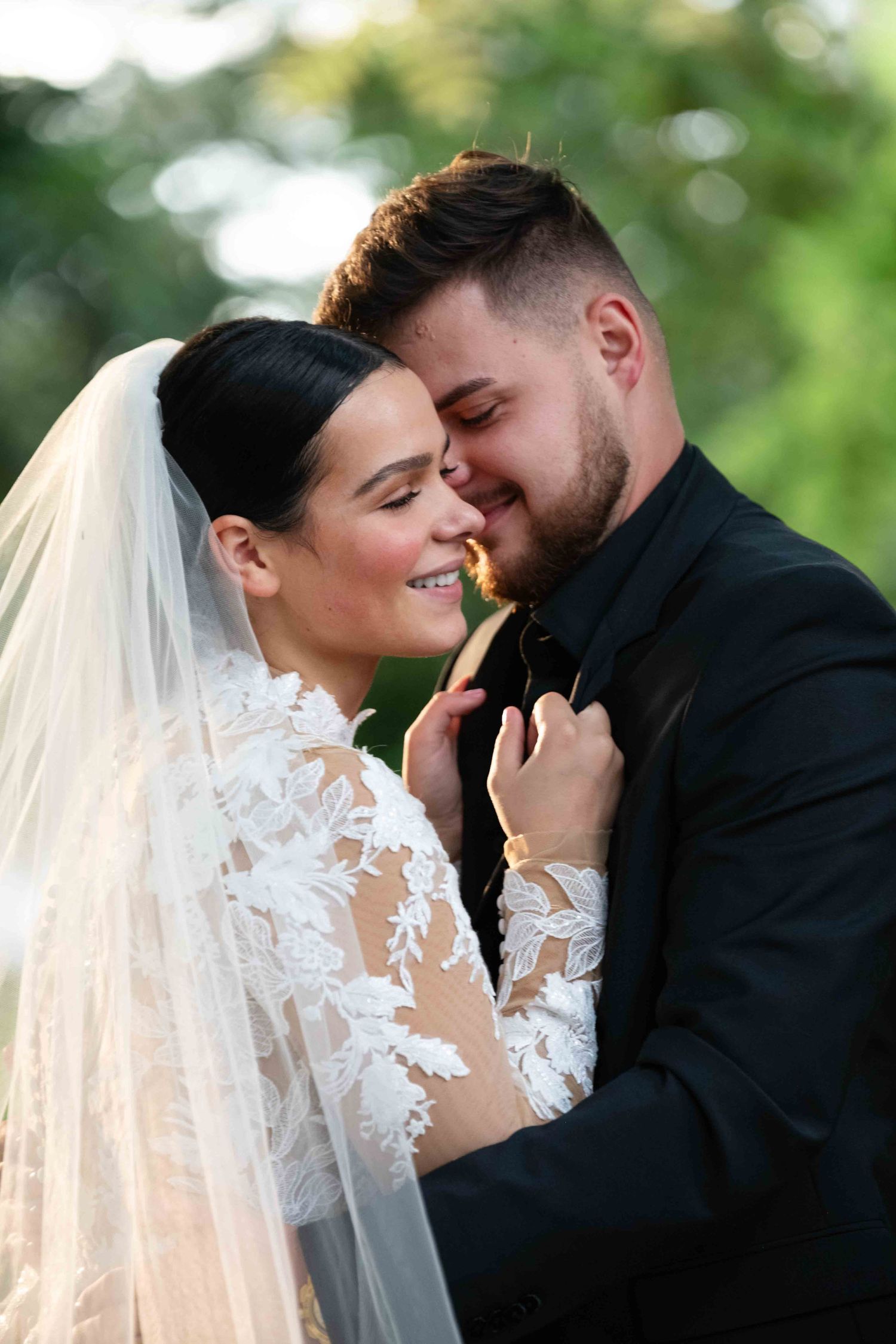 A bride in lace gown and veil shares a tender romantic moment with her groom in a black suit outdoors.