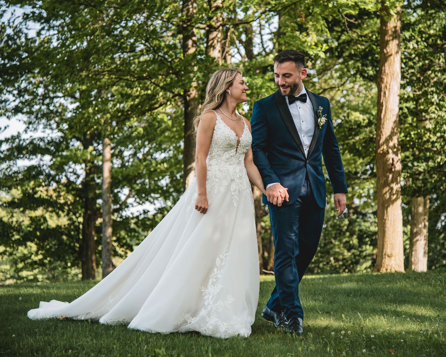 A bride and groom walk hand in hand through a lush green park, smiling at each other on their wedding day.