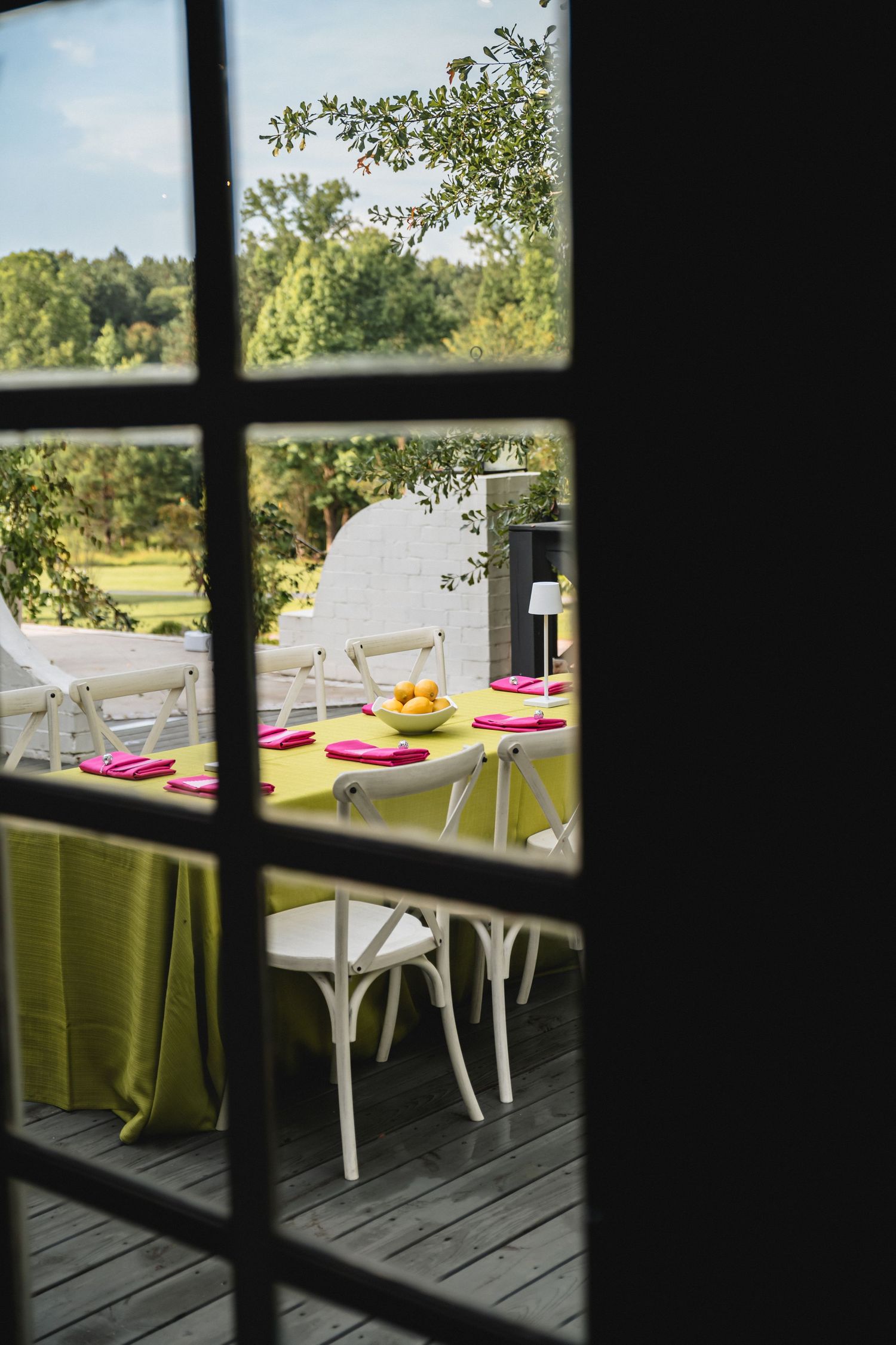 View through window panes of an outdoor reception dining setup with lime green tablecloths and white chairs on a wooden deck.