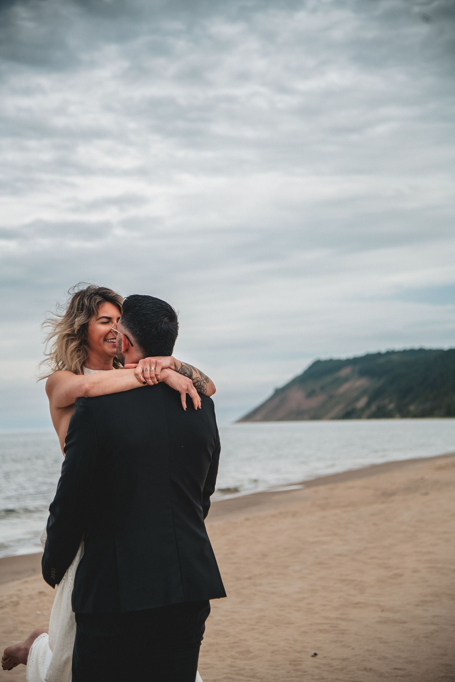 A newlywed couple embraces on a cloudy beach with dramatic overcast skies and coastal bluffs of Sleeping Bear Dunes in the background.