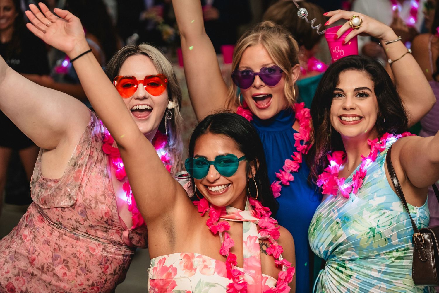 Group of party-goers wearing colorful dresses and sunglasses celebrate with pink leis and raised arms at festive event.