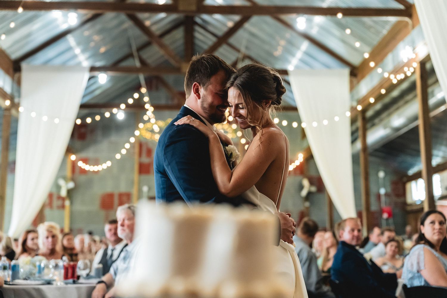 Bride and groom share first dance at rustic barn wedding reception with string lights and guests watching.