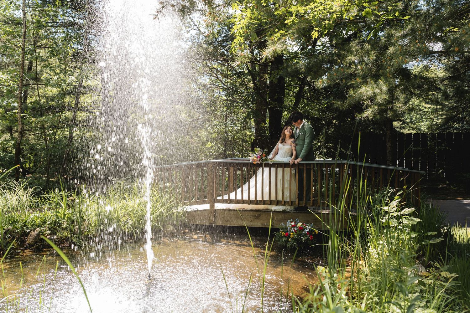 Bride and groom share a romantic moment on a wooden bridge over a pond with a fountain in a lush green garden.