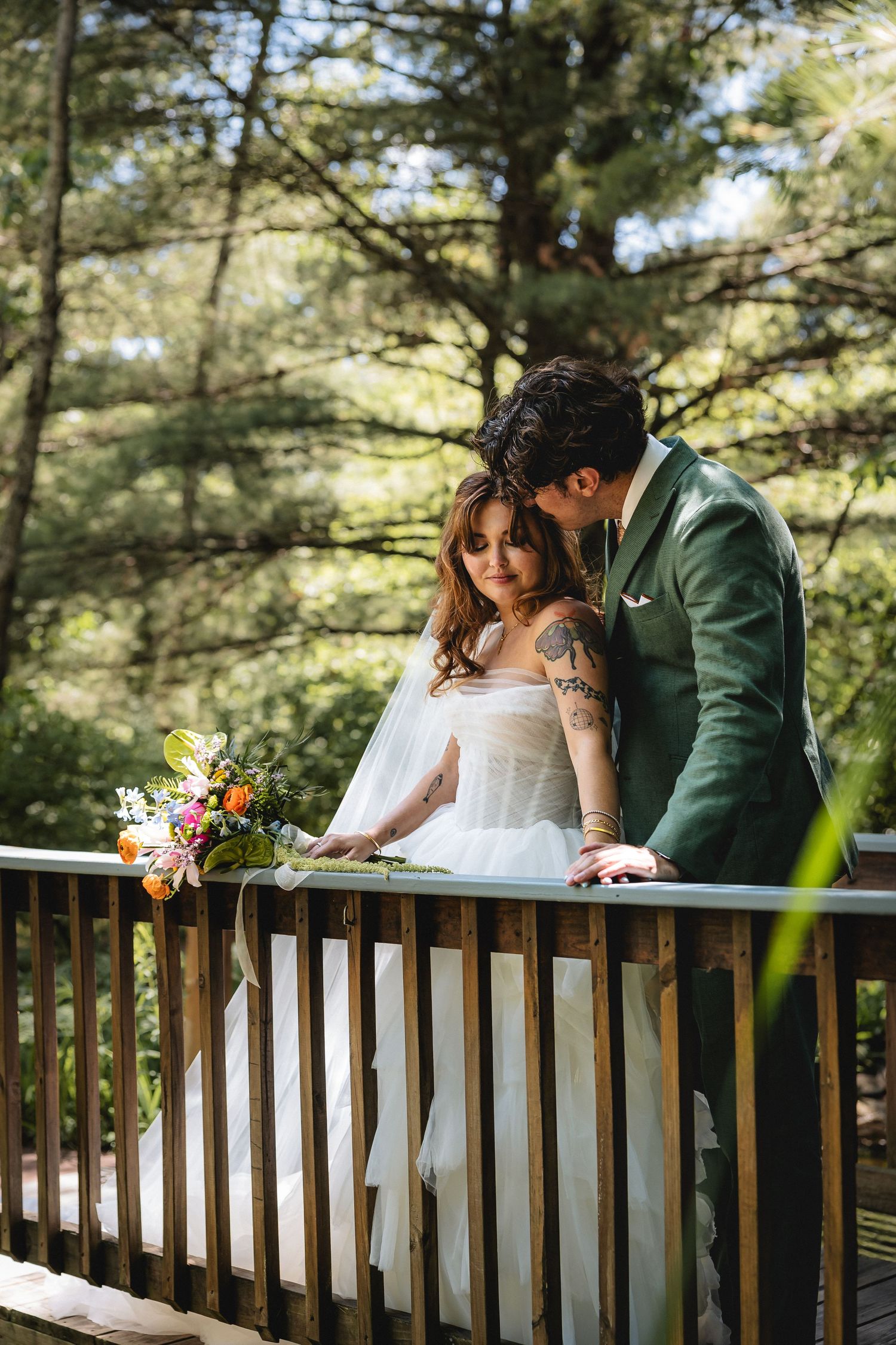 Bride and groom share a romantic moment on a wooden deck surrounded by lush green trees, bride holding colorful bouquet.