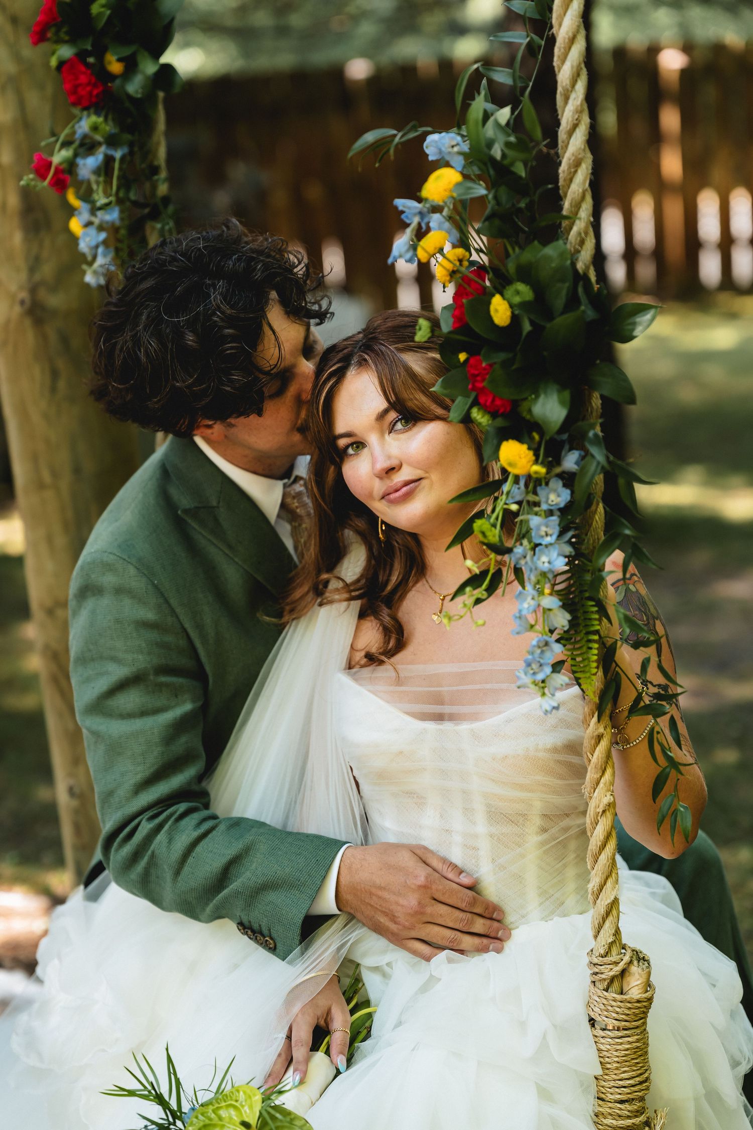 Bride and groom sitting on a floral swing surrounded by colorful blooms in a romantic outdoor wedding setting.