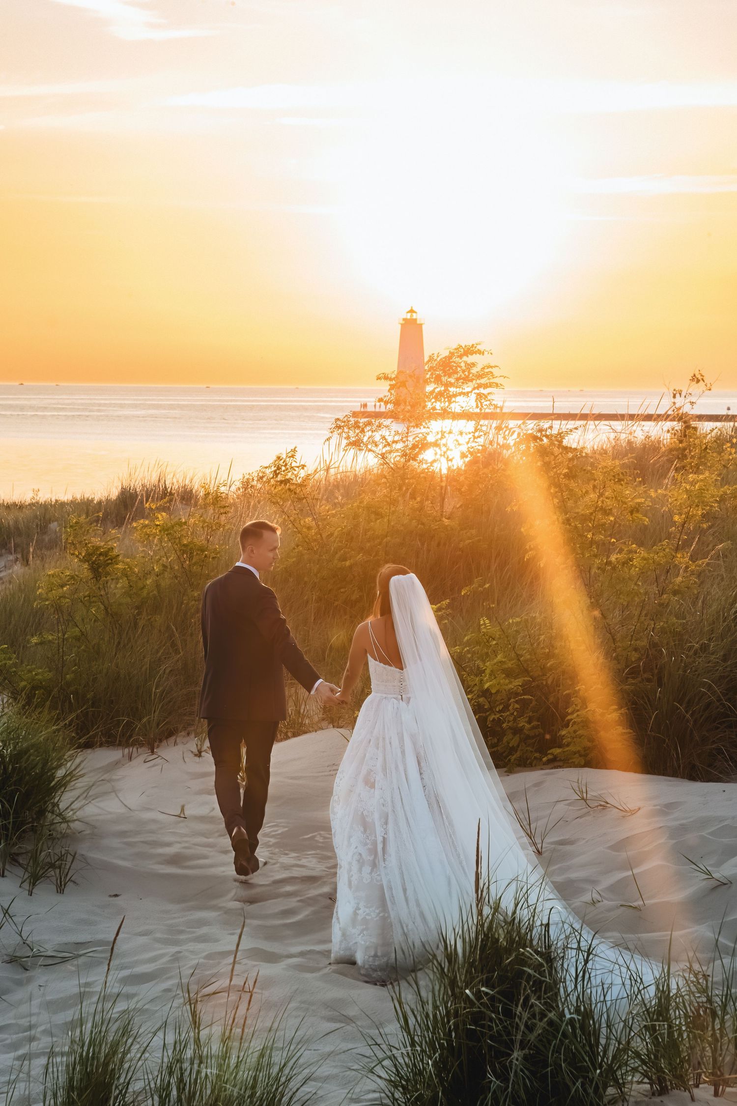 Bride and groom hold hands walking along a sandy dune path toward Frankfort, MI lighthouse and lake at golden sunset.