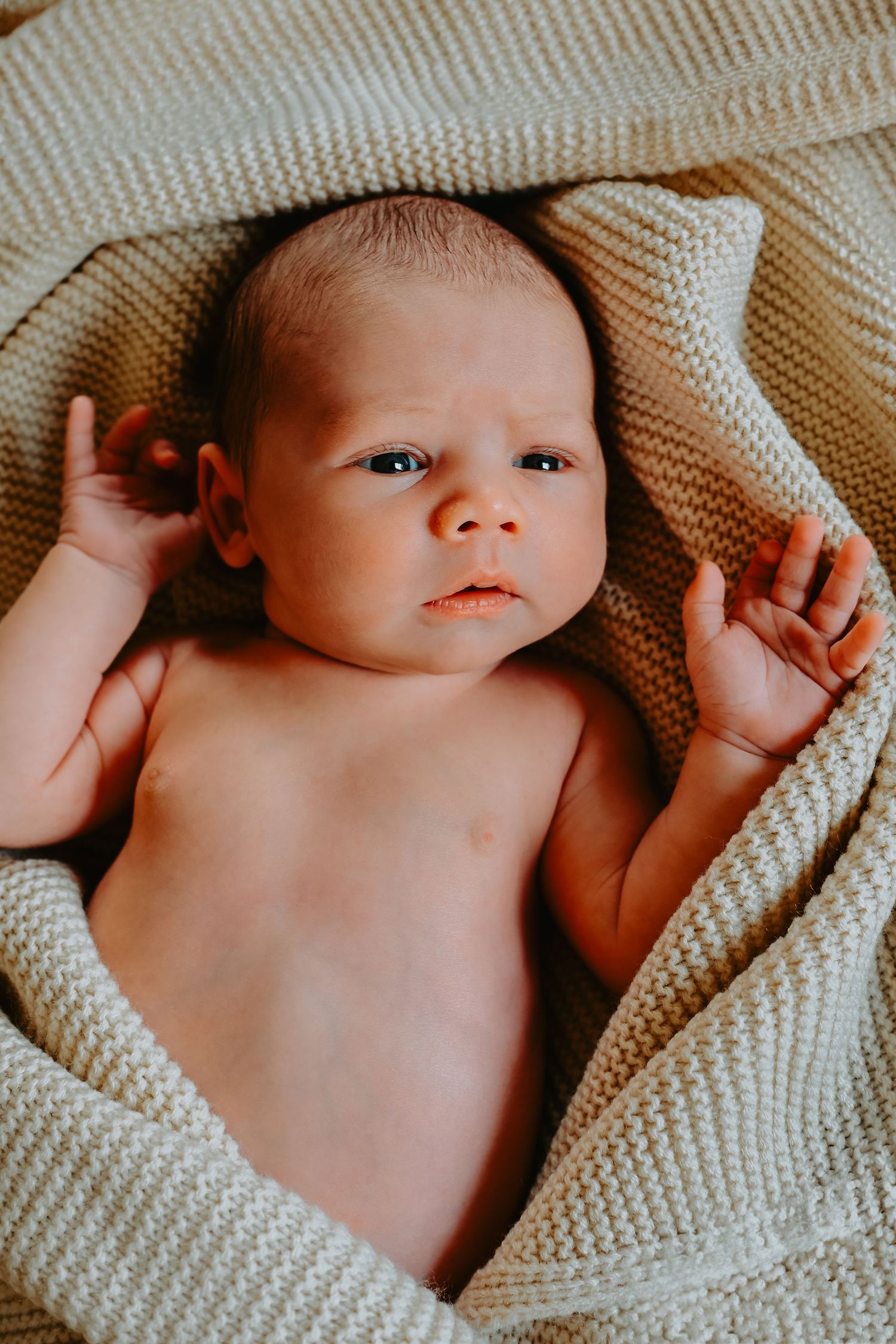 Newborn baby lying on a soft cream knit blanket, looking up with wide eyes and tiny hands raised.