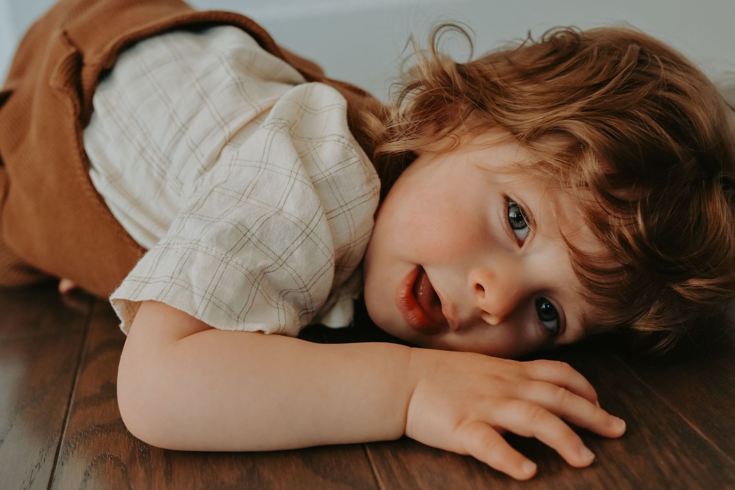 Curly-haired toddler lying on hardwood floor looking up with bright blue eyes, wearing a plaid shirt.