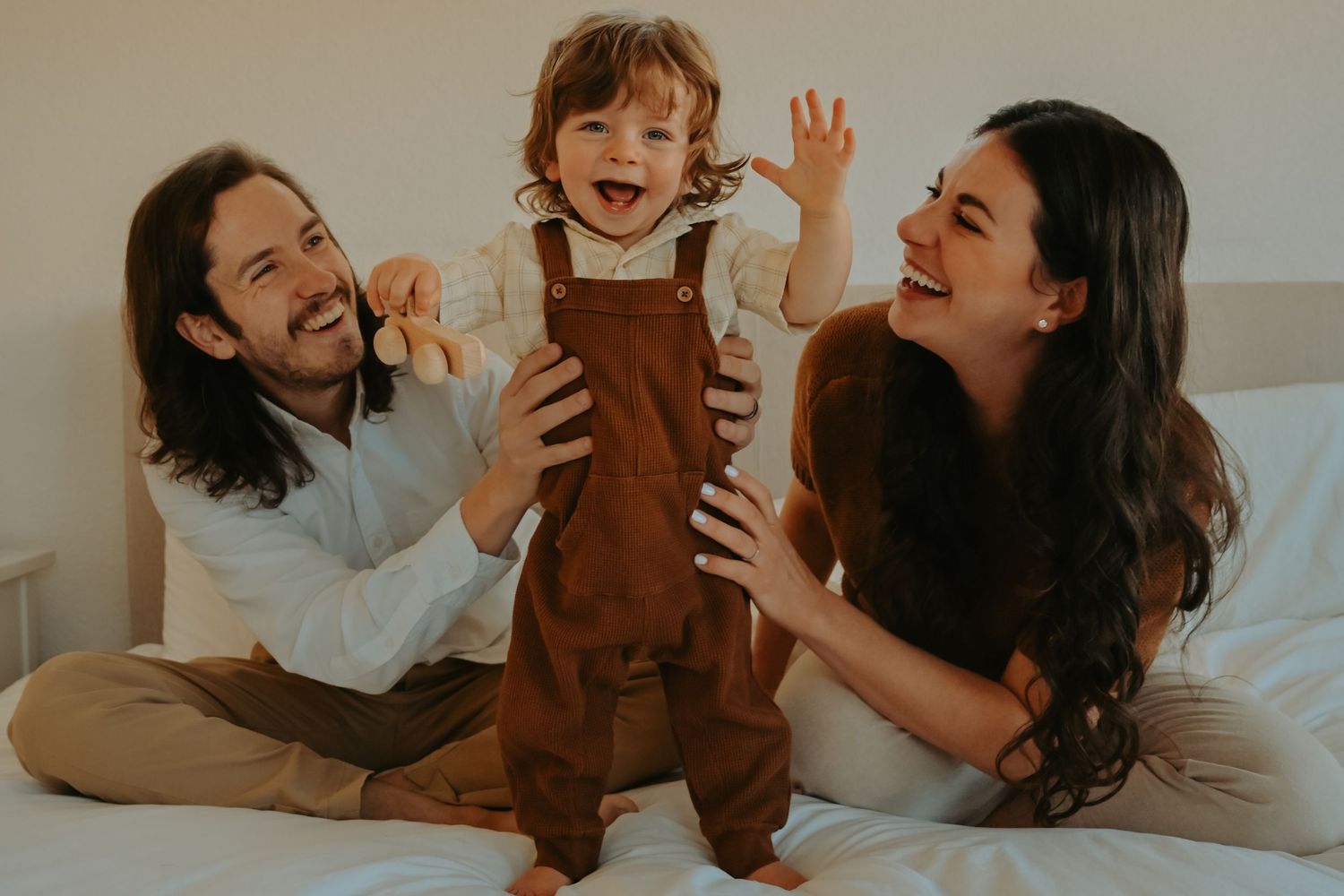 Happy family with parents laughing and holding their joyful toddler on a white bed in a bright room.
