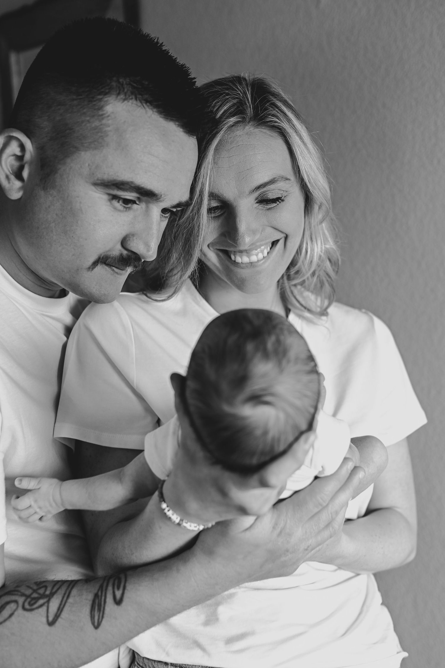 Black and white photo of happy parents in white t-shirts lovingly holding their newborn baby together.