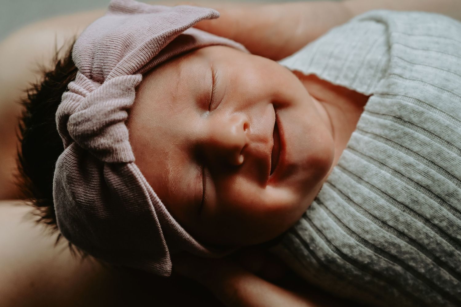 Peaceful newborn baby sleeping wrapped in a gray swaddle with a pink bow hat, cradled in loving arms.